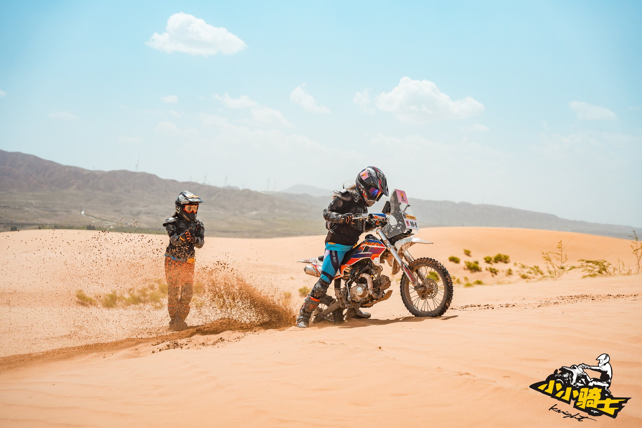 A young rider crossing sandy terrain with coach support nearby.